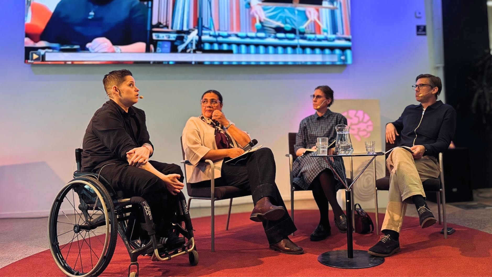 Fyra personer sitter på en scen under ett panelsamtal på Vetenskapsfestivalen. En av deltagarna sitter i rullstol och talar i mikrofon, medan de andra lyssnar. På väggen bakom syns en stor skärm med visuellt material från presentationen.