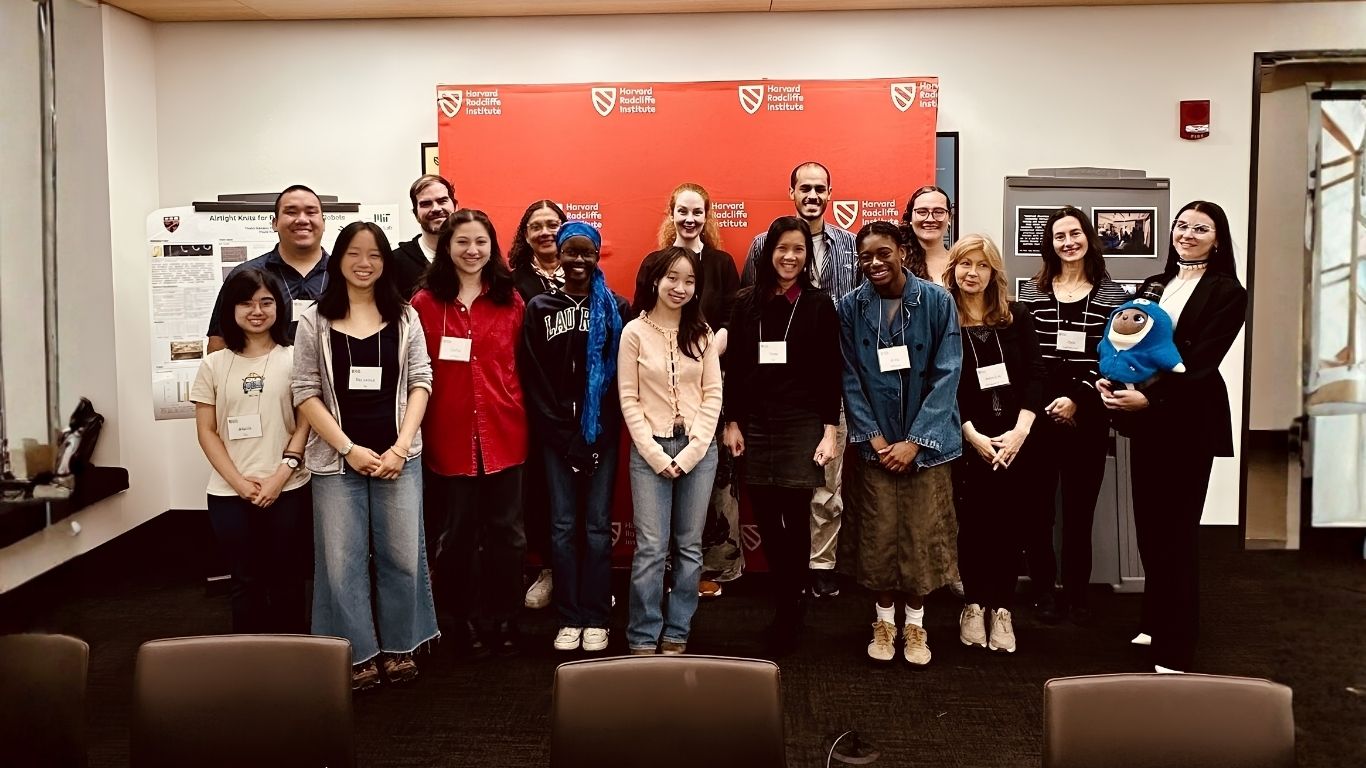 Gruppfoto med flera personer som står tillsammans framför en röd vägg med Harvard Radcliffe Institutes logotyp. Personerna ler mot kameran och bär namnskyltar. Bilden är tagen inomhus i ett konferensrum.