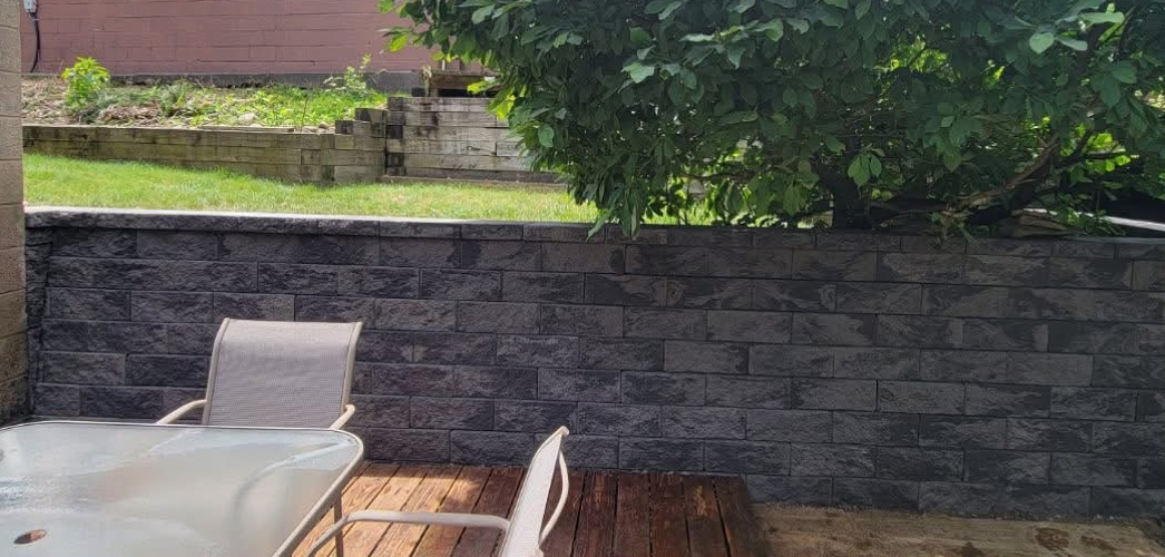 Backyard patio with glass table, two white mesh chairs, wooden deck, and a dark stone retaining wall with grass and tree above.