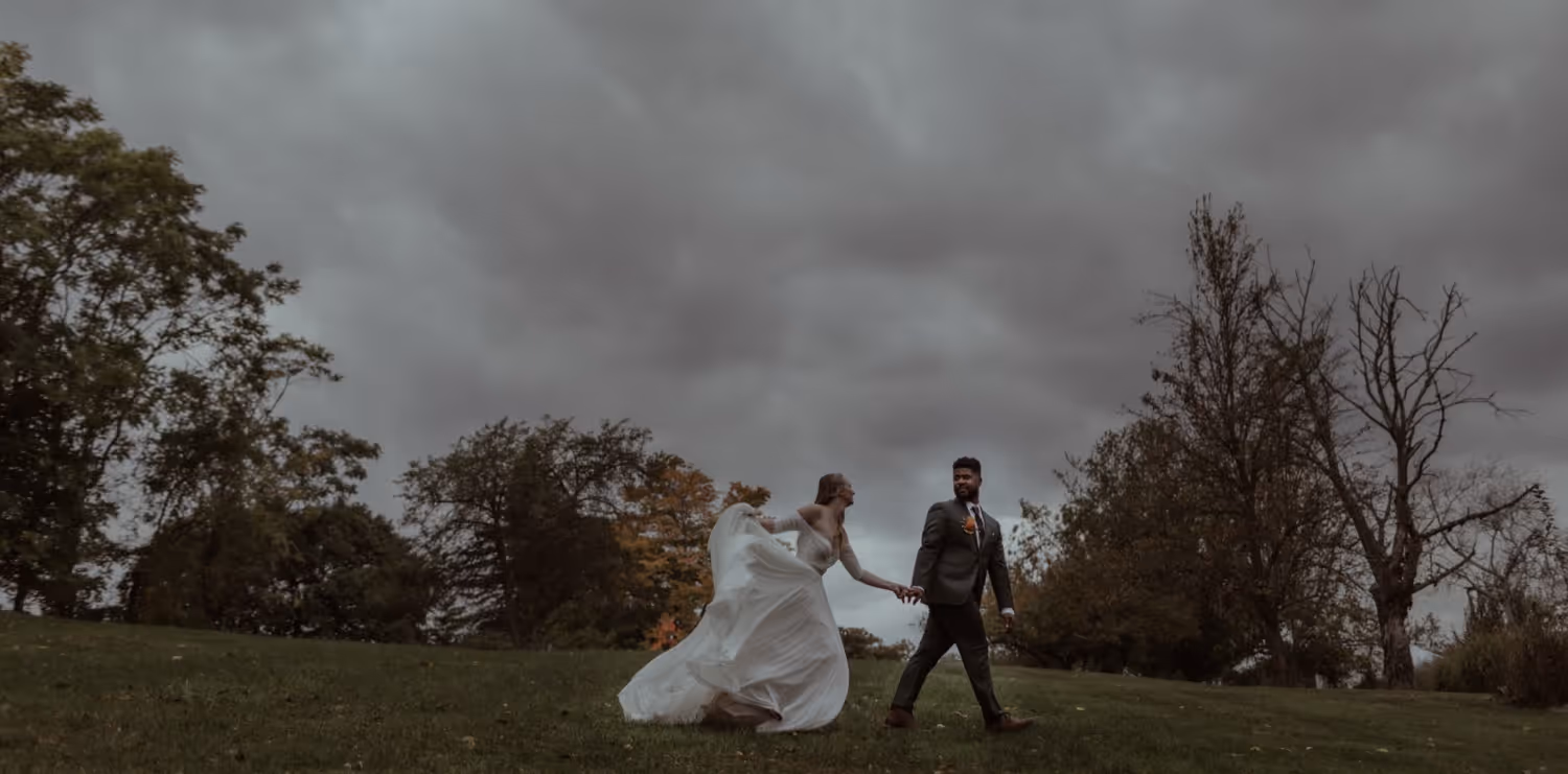 Bride in white gown and groom in dark suit holding hands and walking on grass under cloudy sky with trees around.