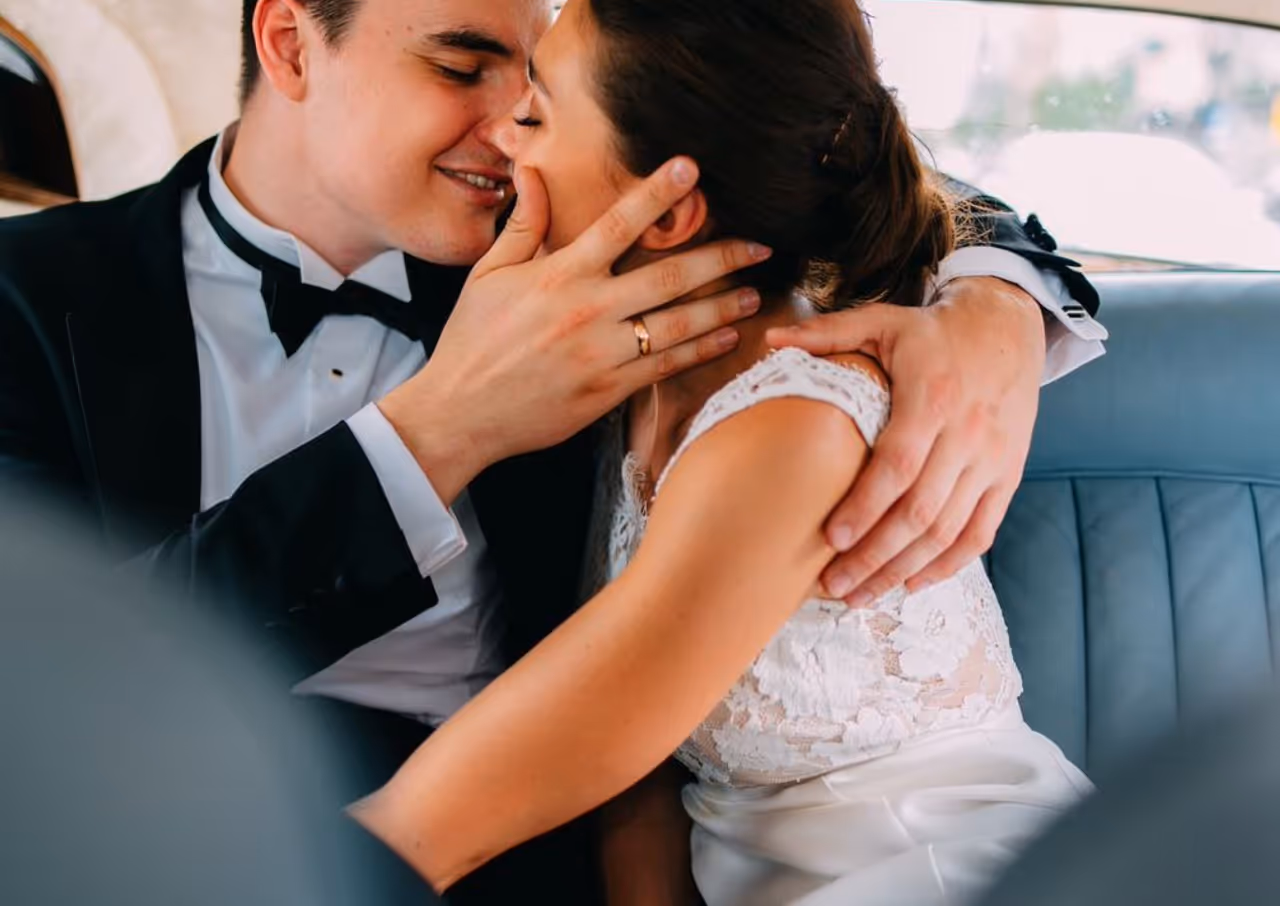 Bride and groom embracing closely inside a vehicle, the groom gently holding the bride's face and the bride wearing a lace wedding dress.