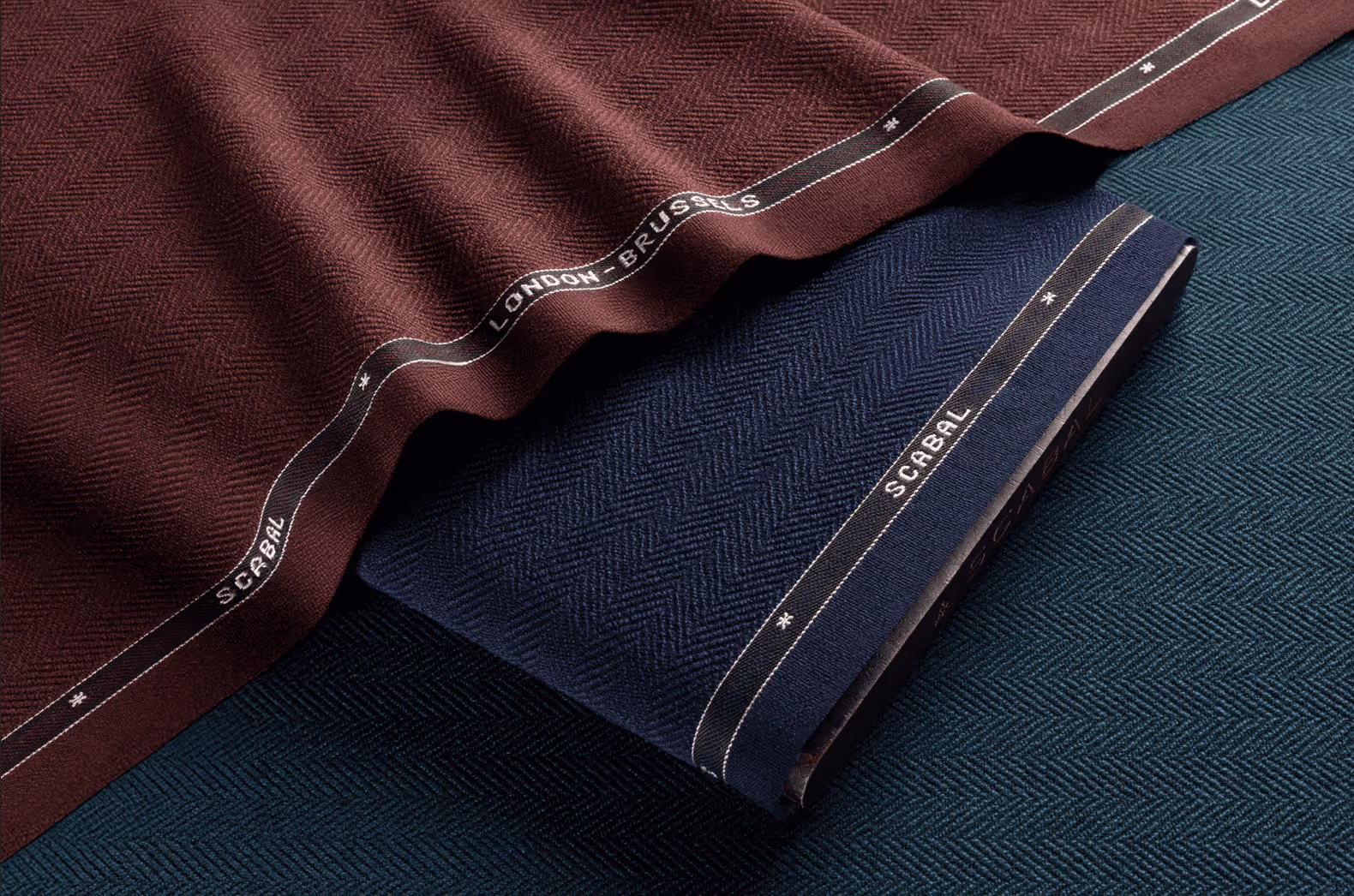 Folded wool fabrics in burgundy, navy, and teal with herringbone patterns and SCRABAL branding on the edges.