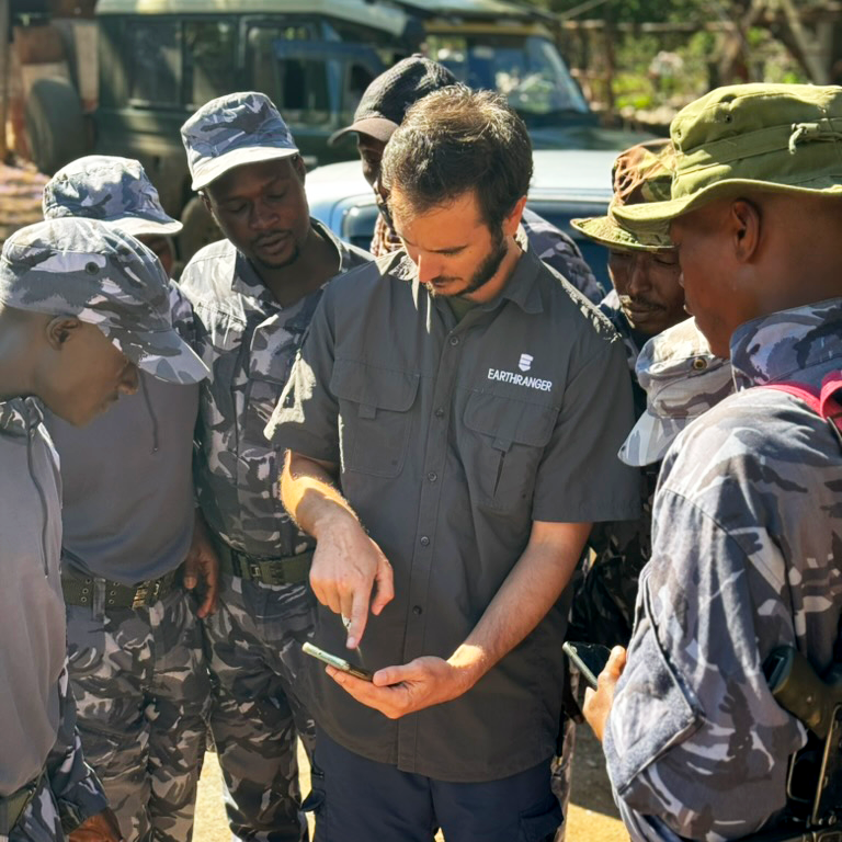 A man wearing a shirt with the EarthRanger logo on it points at a mobile phone, which men in ranger uniforms observe the screen.