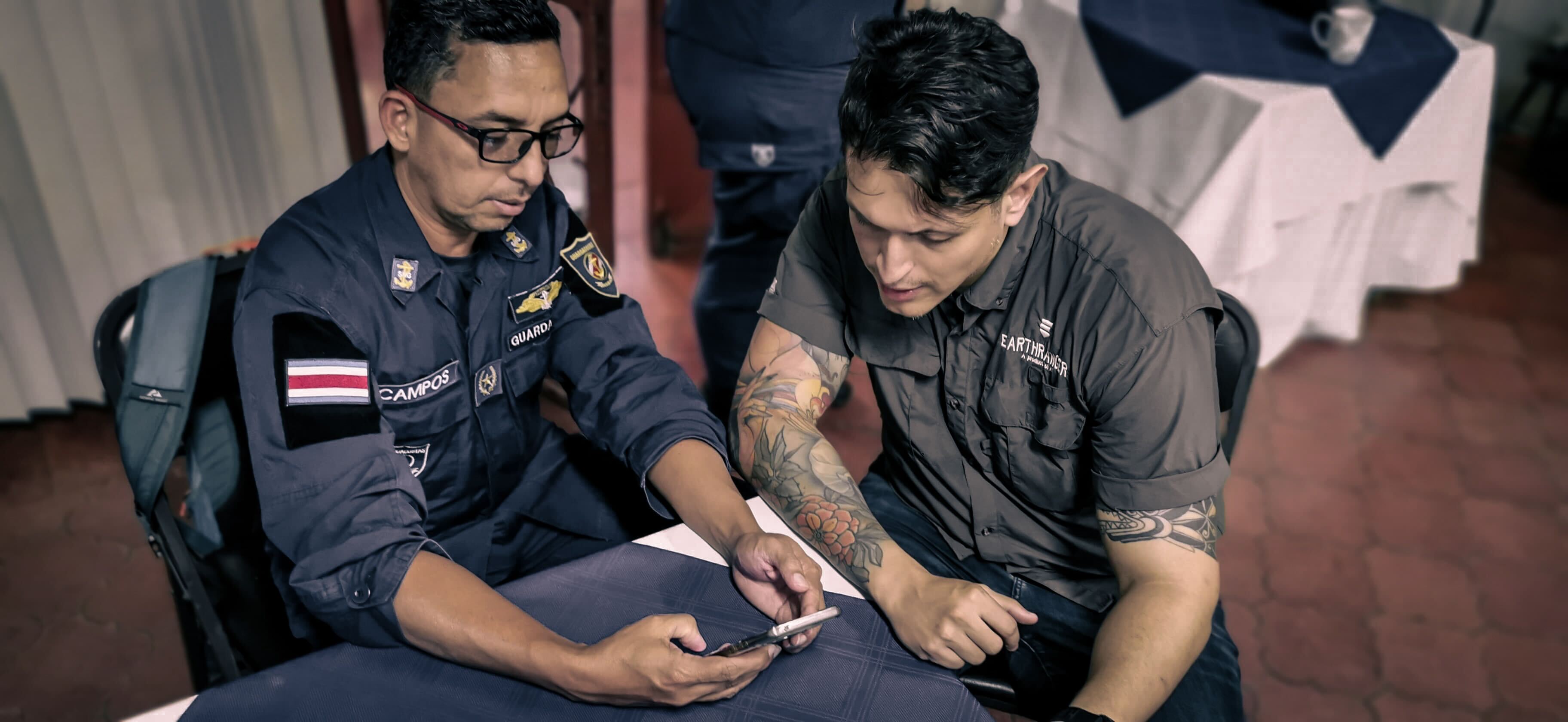 Two men sit together looking at a phone. One man is wearing a ranger's uniform with a Costa Rican flag on it. The other man is wearing a button up shirt with the EarthRanger logo on it.