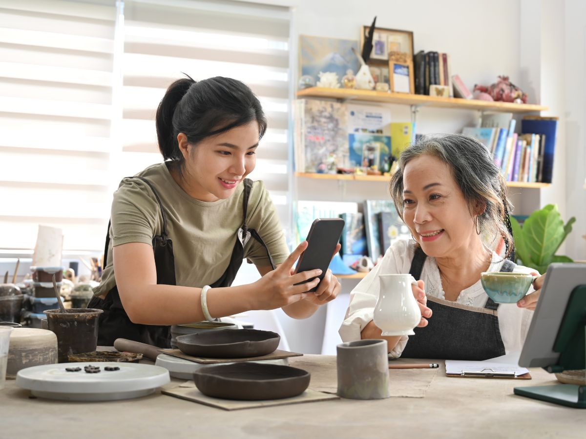 A mother and daughter team taking product photos of their ceramic products in a bright and airy workshop. The daughter holds her phone to eye level to take a photo of a white ceramic jug as her mum holds it for her.