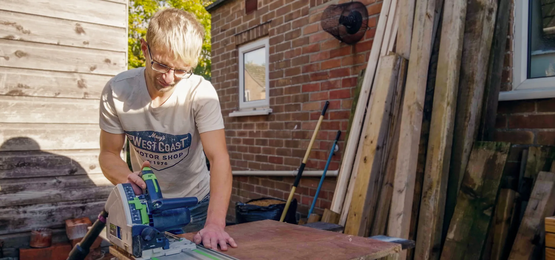 Man with glasses using a power saw to cut a wooden board outdoors beside a brick wall and stacked wood.