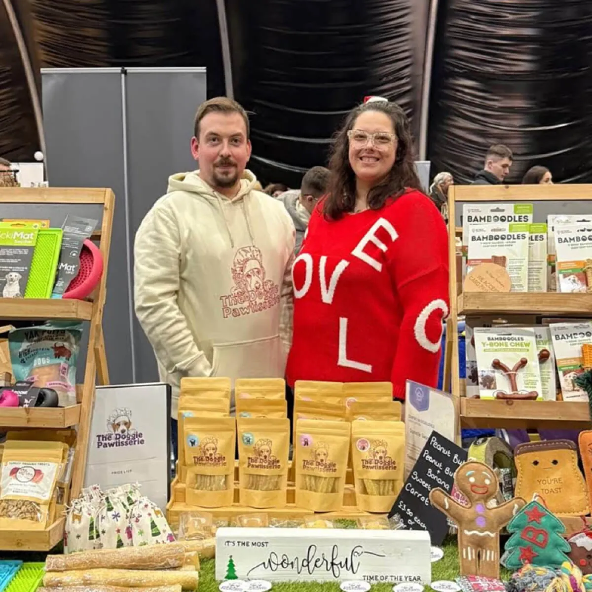 Two vendors standing behind a table filled with dog treats and toys, including packaged snacks and plush toys, at a market stall.