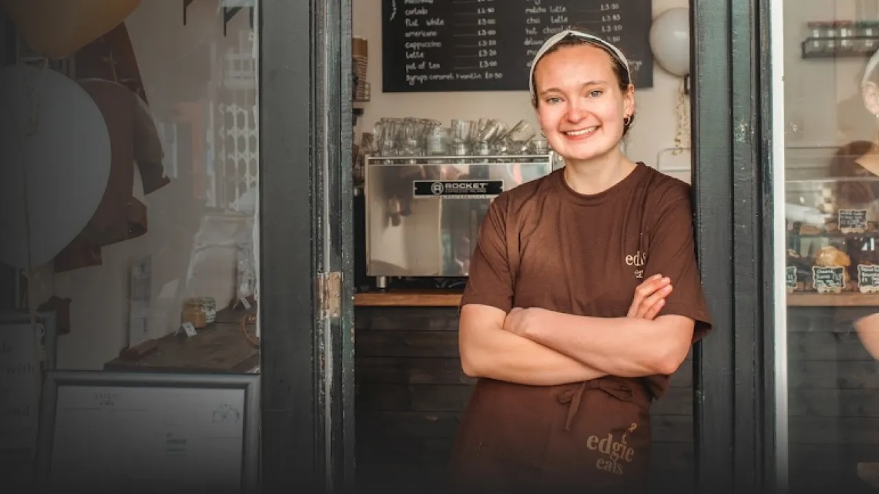 Smiling barista standing with crossed arms in a coffee shop doorway wearing a brown edgie eats uniform.
