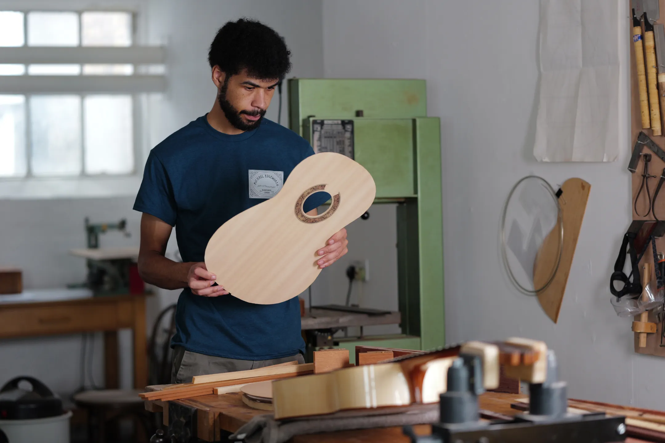 Man in blue shirt inspecting a wooden guitar body in a woodworking workshop.