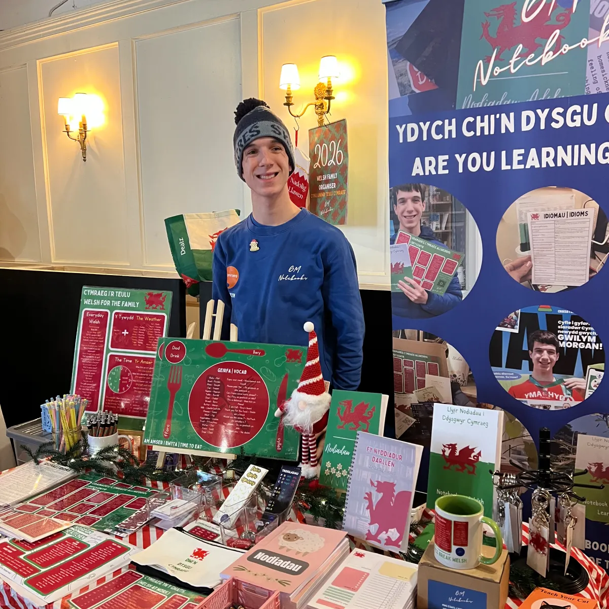 Young man in a blue sweatshirt and wool hat standing behind a table displaying Welsh language learning materials and merchandise.