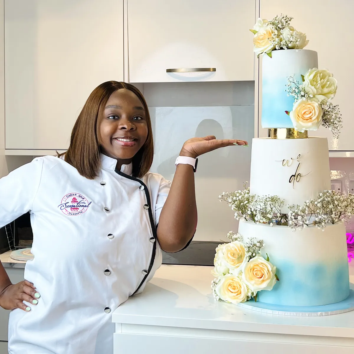 Female baker in white uniform presenting a three-tiered white and blue wedding cake decorated with white and yellow roses and flowers.