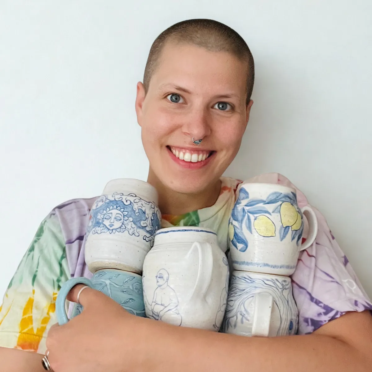 Smiling person with shaved head holding six decorated ceramic mugs featuring blue and yellow designs.