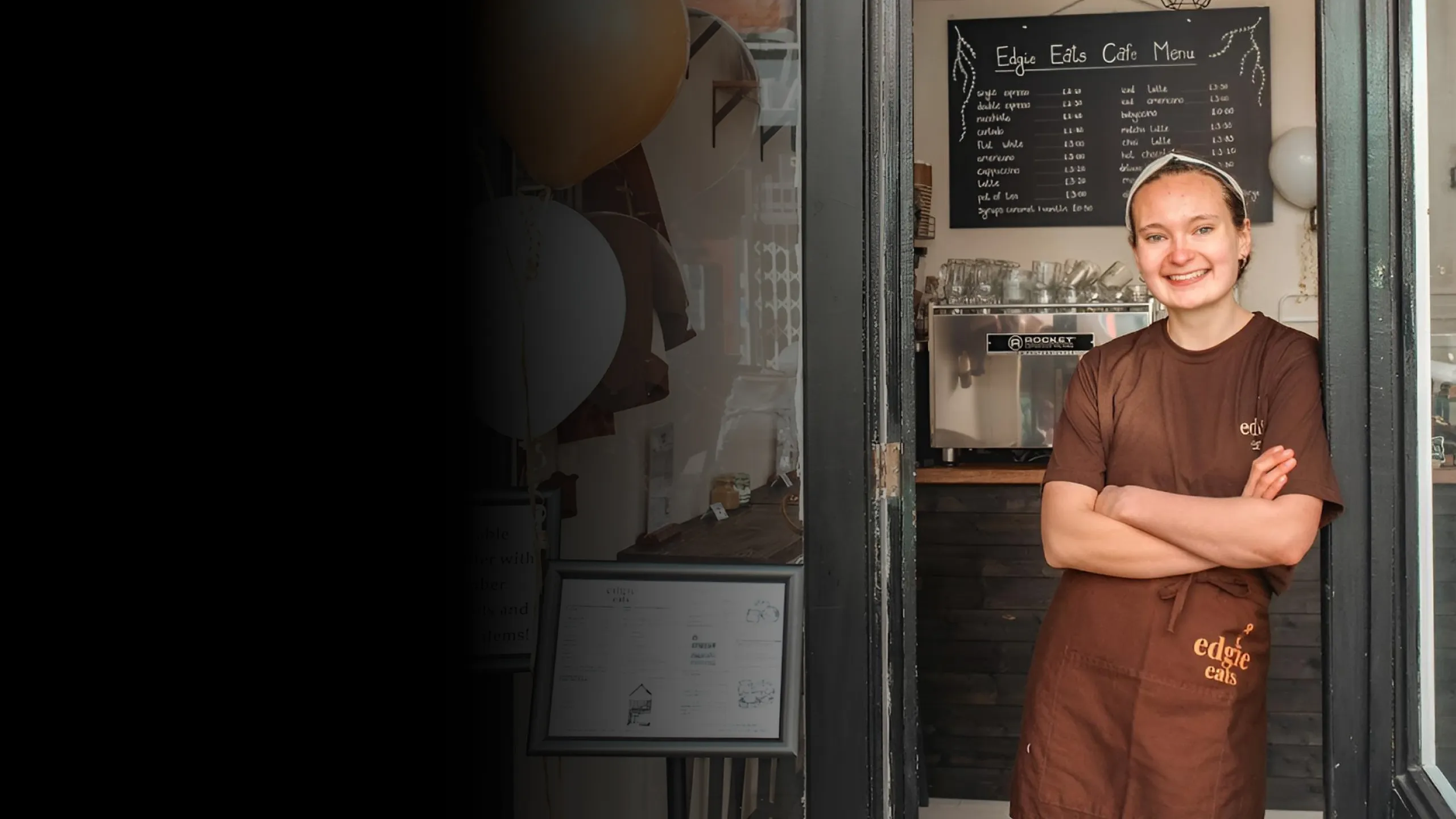 Smiling woman wearing a brown Edgie Eats apron and shirt standing with arms crossed in a cafe doorway.