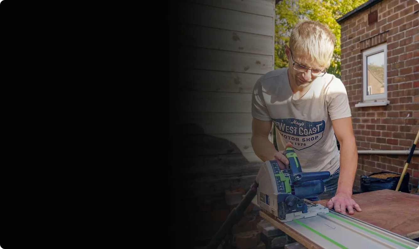 Young man with glasses using a track saw to cut a wooden board outdoors next to a brick building.