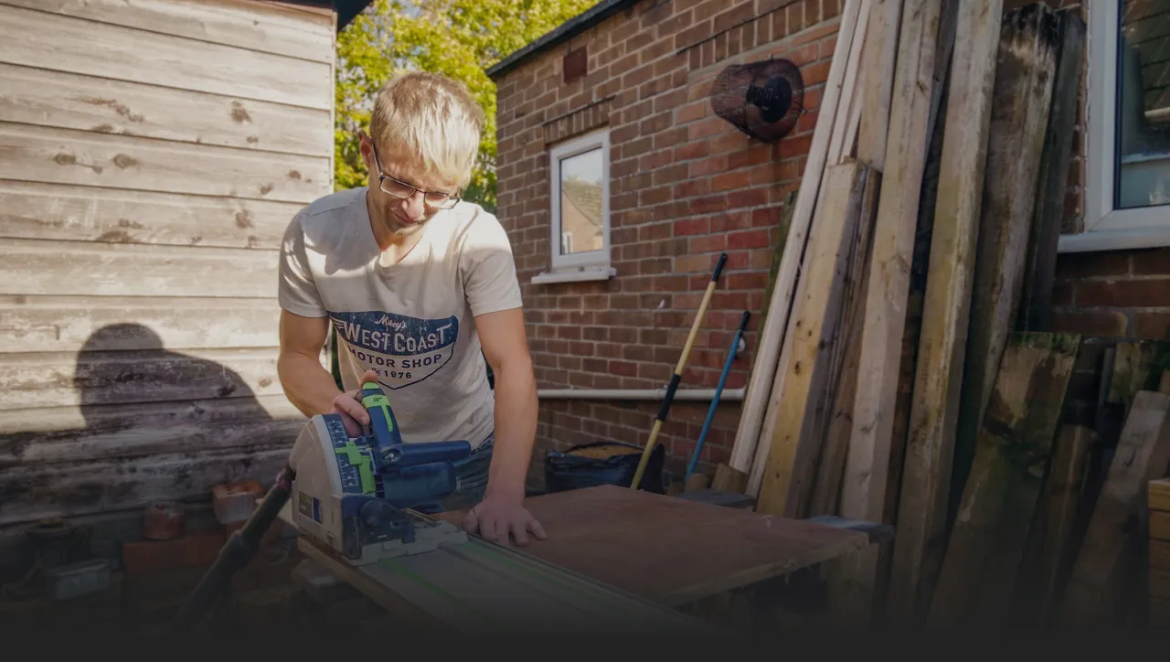 Young man with glasses cutting a wooden board with a track saw outdoors, beside a brick wall and stacked timber.