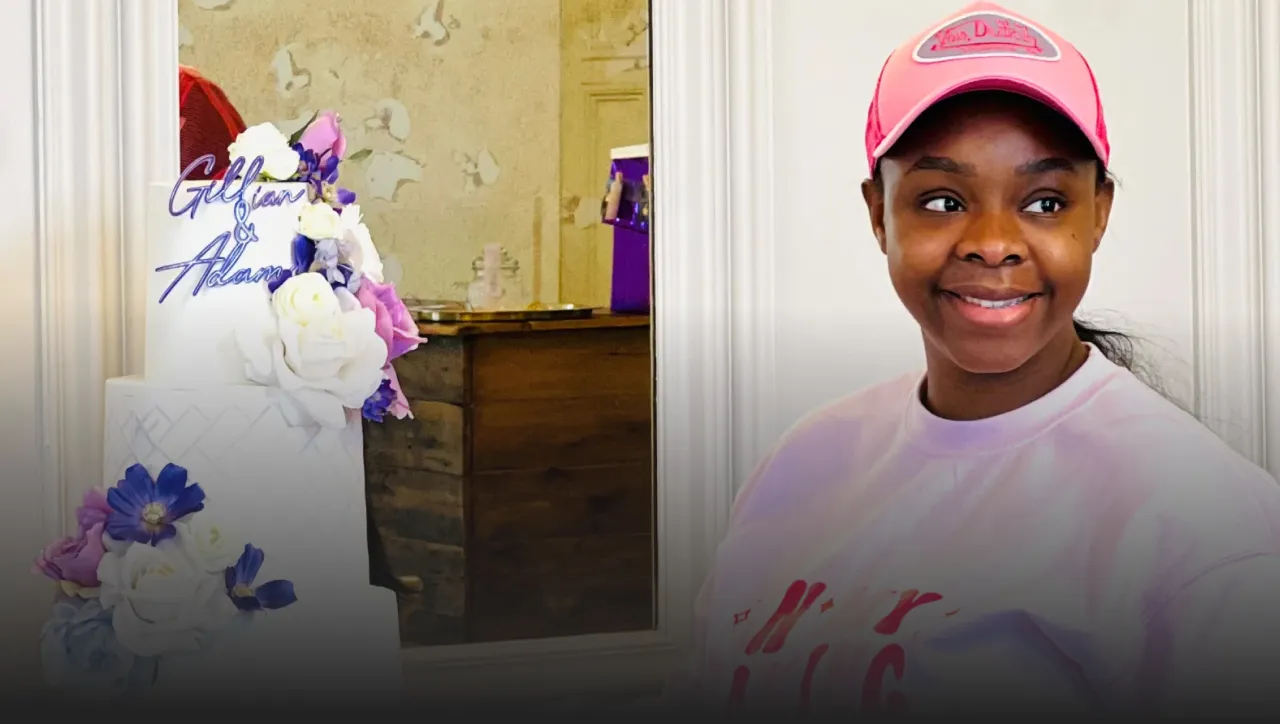 Smiling young woman in a pink cap and pink sweatshirt standing next to a tiered cake decorated with purple and white flowers.