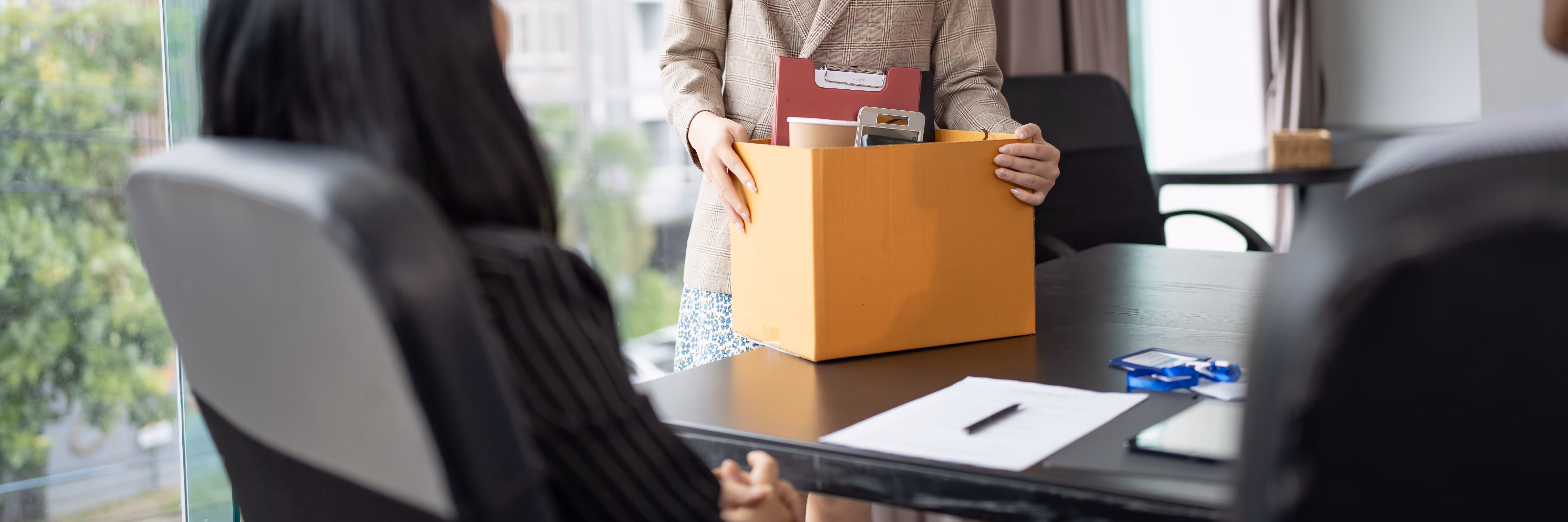 Eine Arbeitnehmerin packt ihre Unterlagen zusammen. Eine weitere weibliche Person sitzt an ihrem Schreibtisch.