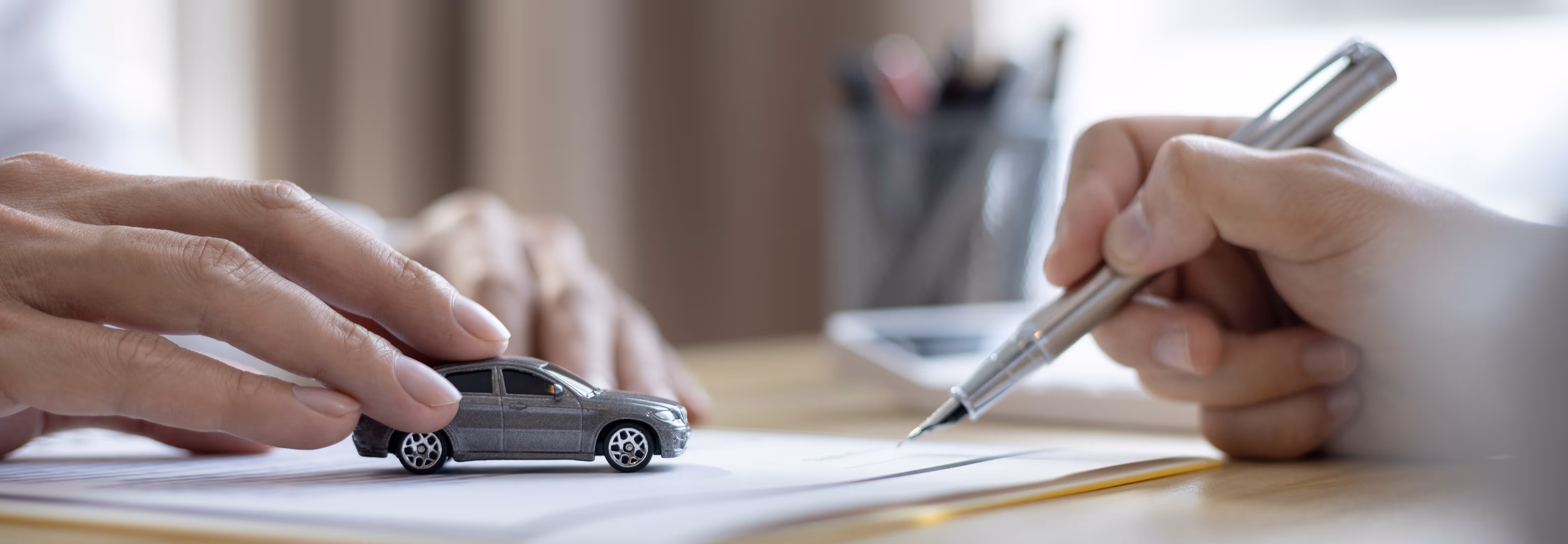 Eine Frau legt ihre Hand auf ein Spielzeugauto, dass auf einem Stapel Dokumente liegt. Ihr gegenüber hält ein Mann einen Stift in der Hand.