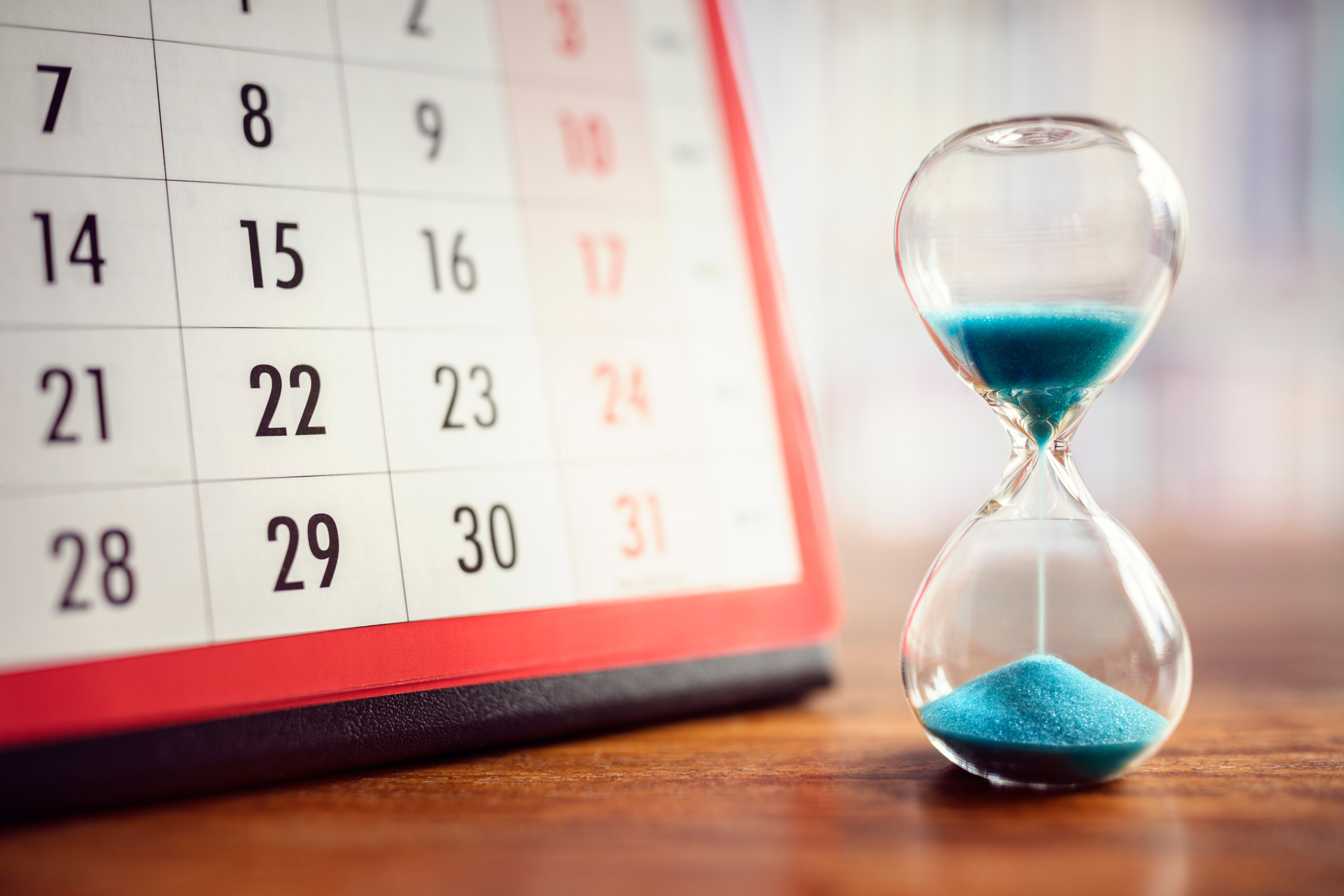 Eine gläserne Sanduhr mit blauen Sand neben einem Kalender auf einem Holztisch.