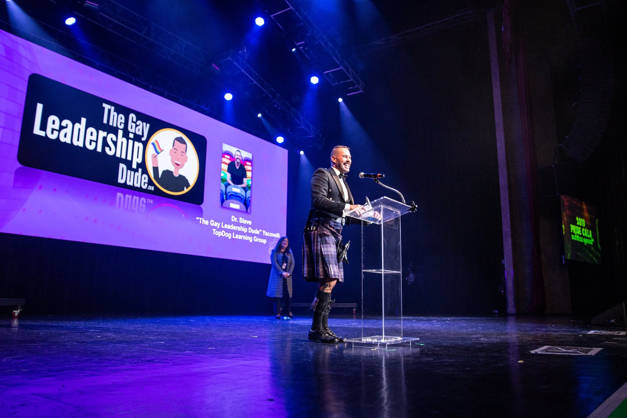 White male standing on a stage, speaking into microphone, with a huge screen behind him with title "The Gay Leadership Dude."