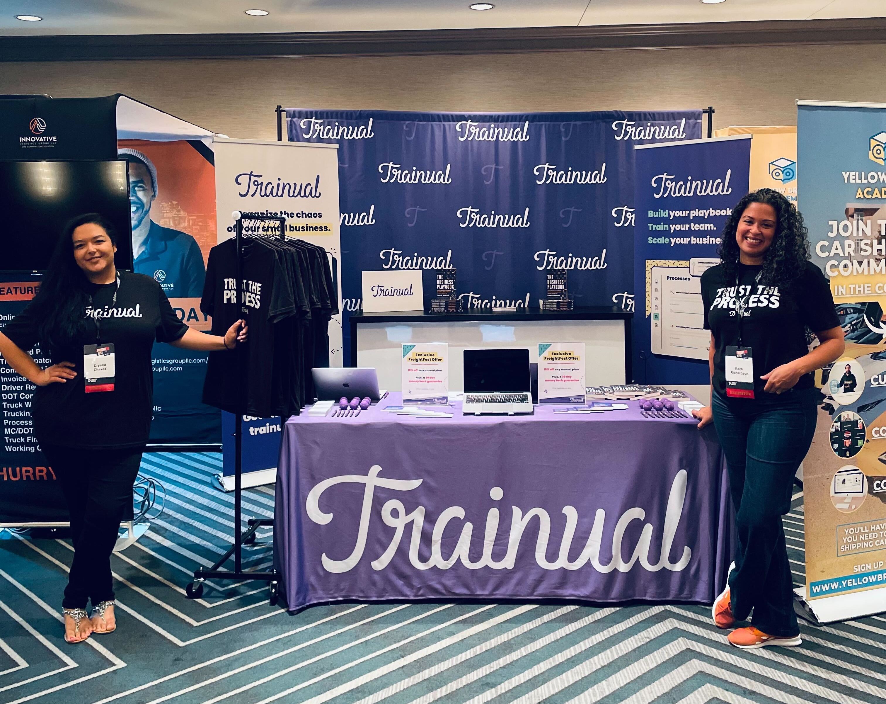 Two women posing in front of a "Trainual" conference booth.