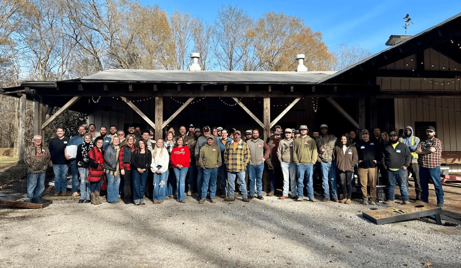 Group photo of about 45 people standing outside in front of a wooden pavilion on a sunny day with bare trees in the background.