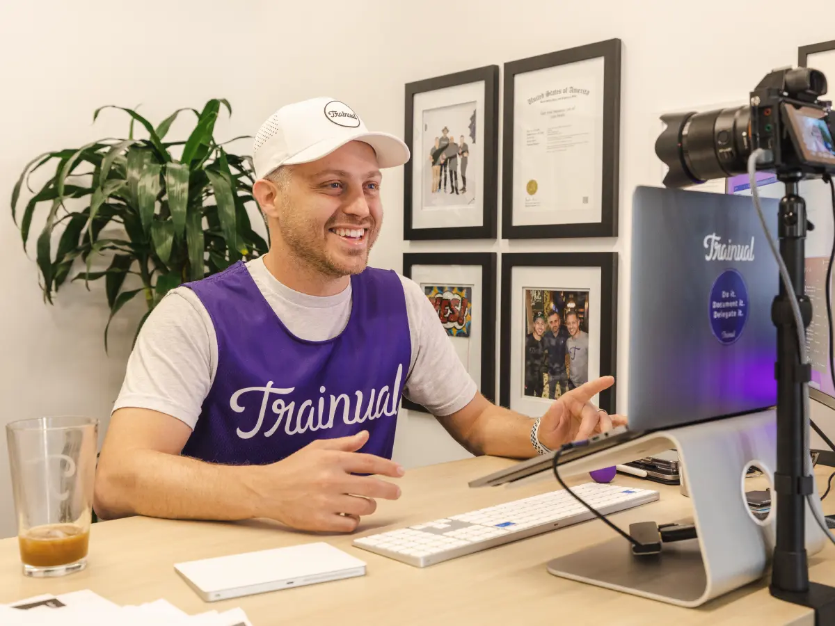Smiling man wearing a Trainual cap and vest sitting at a desk, engaging with a camera and computer setup.