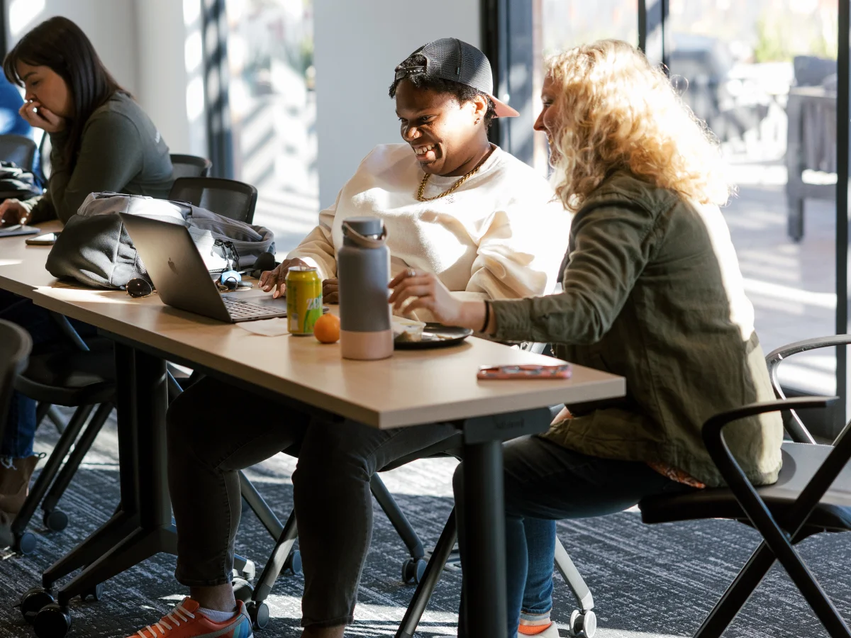 Two people sitting at a desk sharing a laugh while working on a laptop in a bright office space.