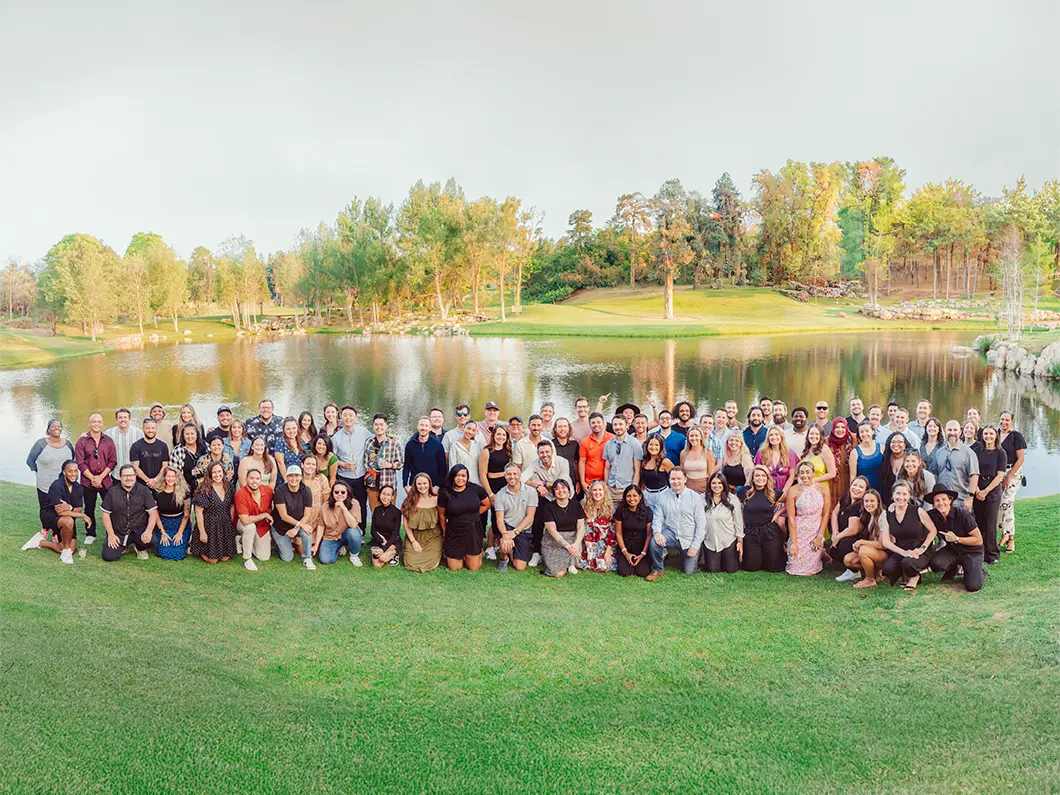 Large diverse group of people posing together on green grass near a lake with trees and hills in the background.