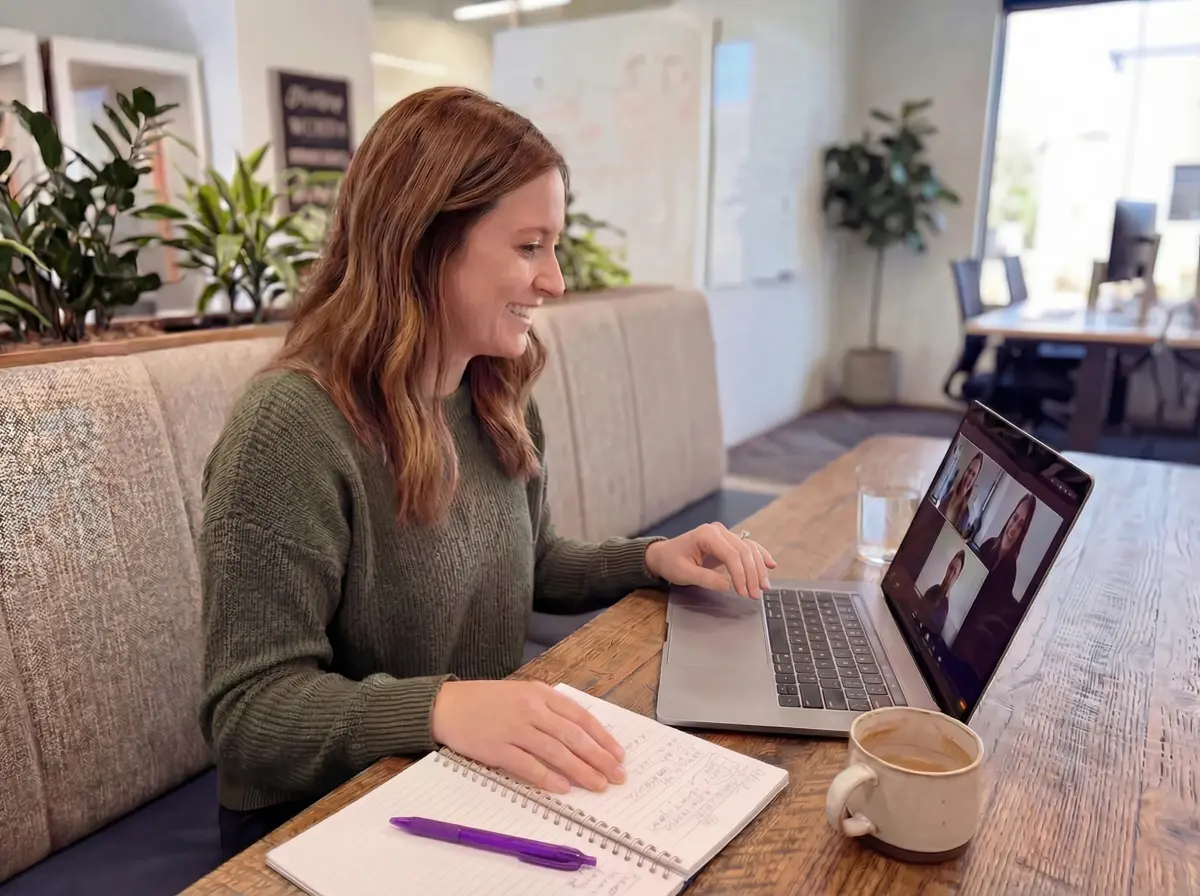 Woman in green sweater sitting at a wooden table, video conferencing on a laptop with four people visible on screen, with a notebook, pen, coffee cup, and glass of water nearby.