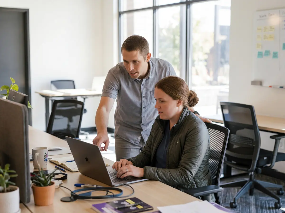 Man standing and pointing at laptop screen while woman seated typing in a modern office workspace.