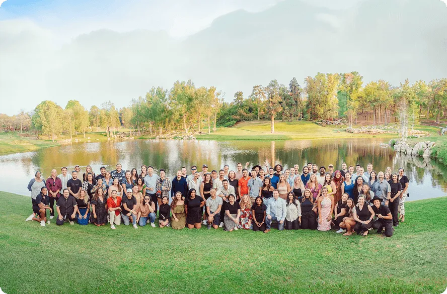 Large group of diverse people posing outdoors on green grass near a pond with trees and blue sky.