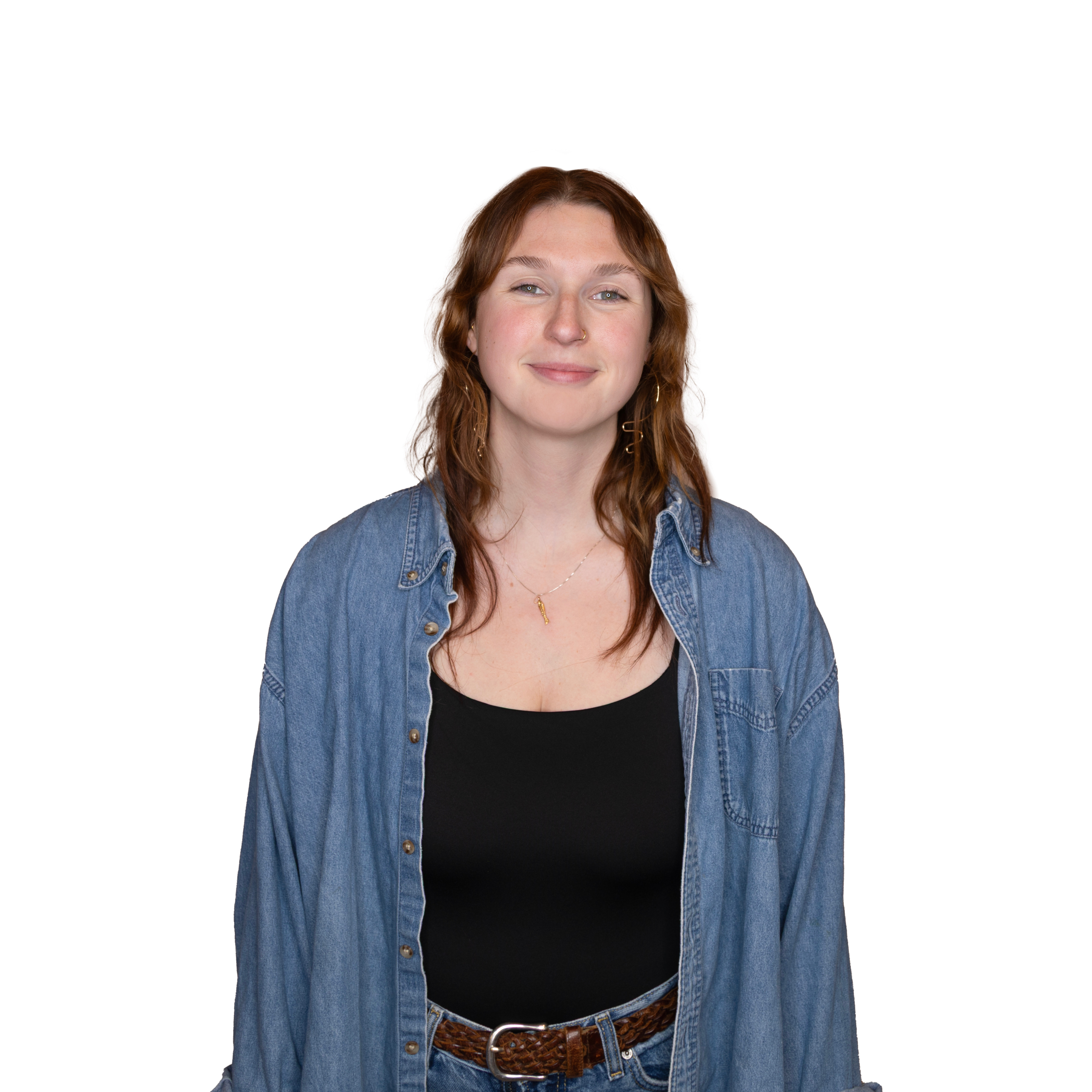 Smiling woman with wavy brown hair wearing a black top, denim shirt, jeans, and a brown braided belt against a white background.