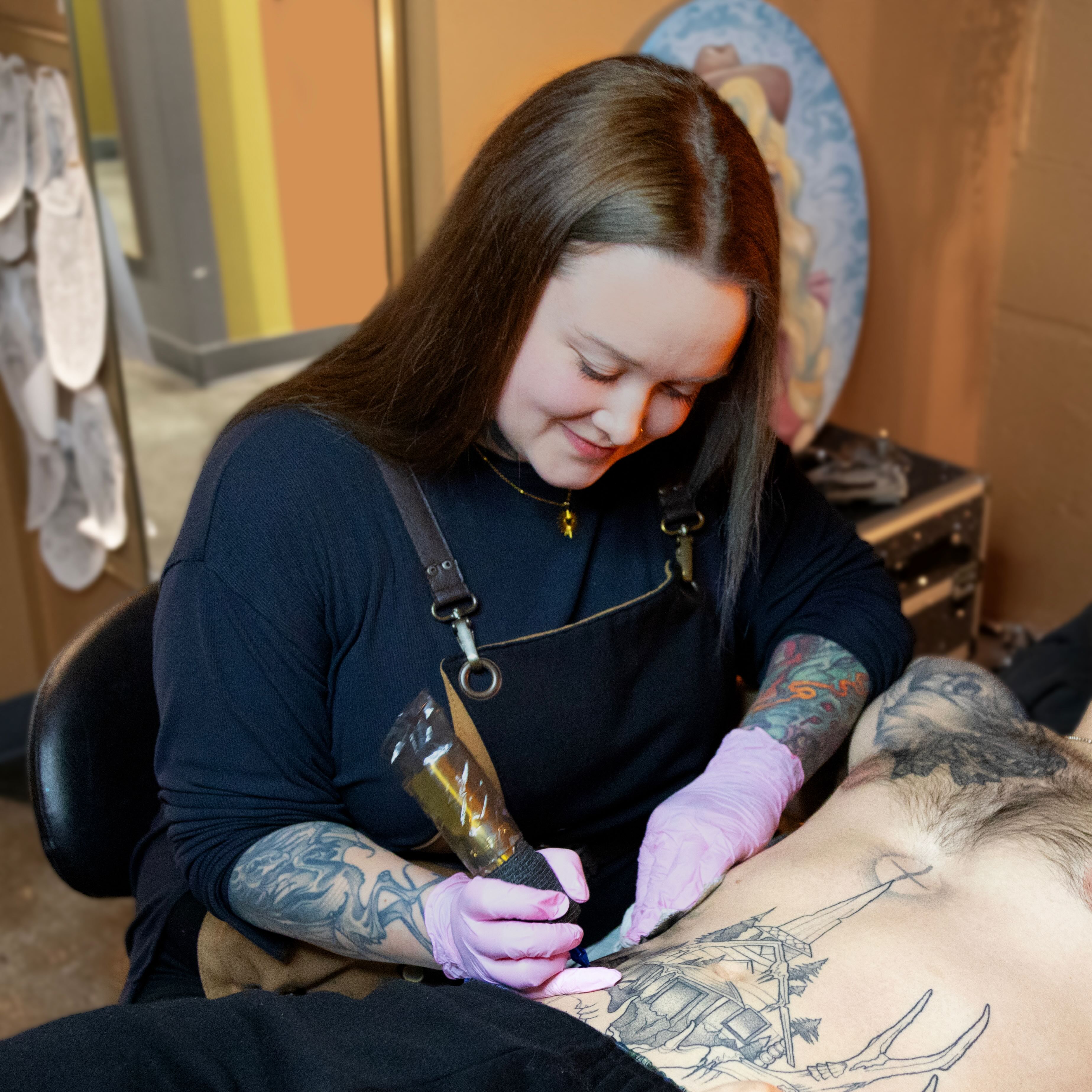 Tattoo artist with long brown hair and sleeve tattoos working on a man's large chest tattoo in a shop.