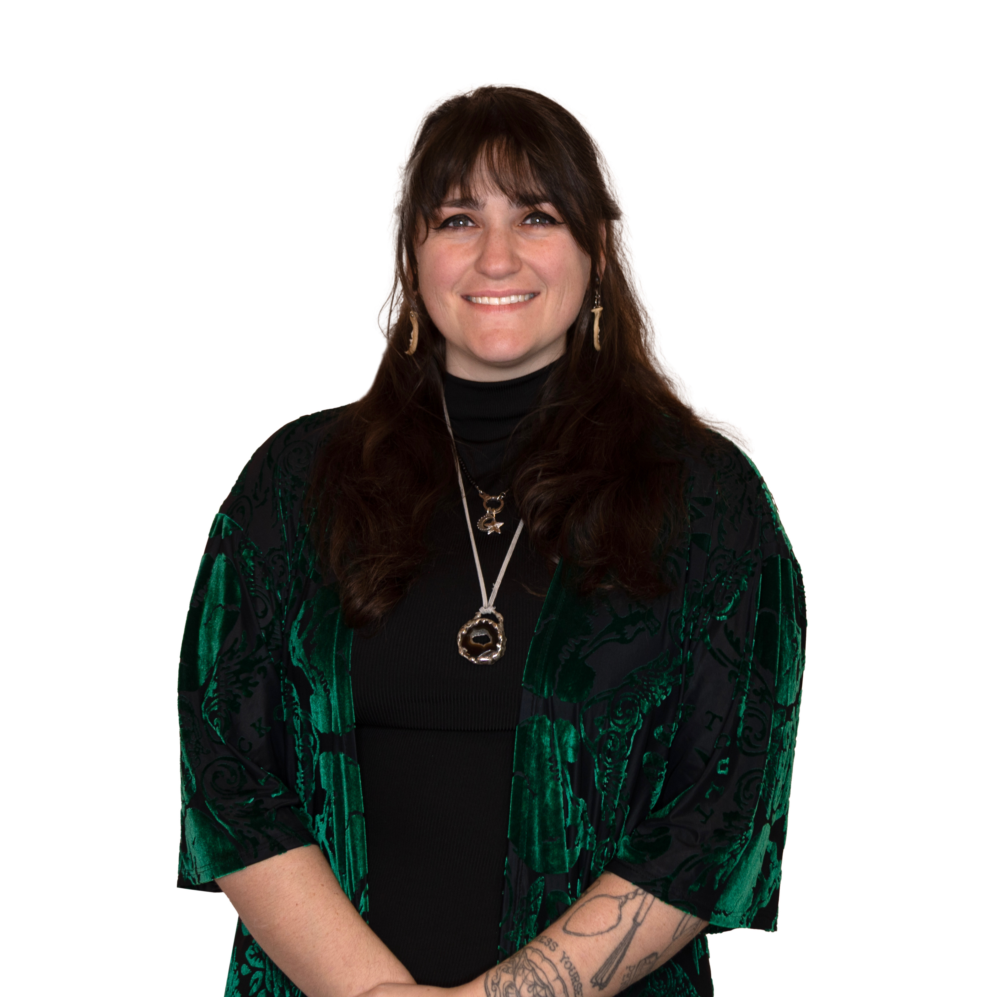 Smiling woman with long dark hair wearing a dark green patterned kimono over a black top, with tattoos on her forearm and layered necklaces.