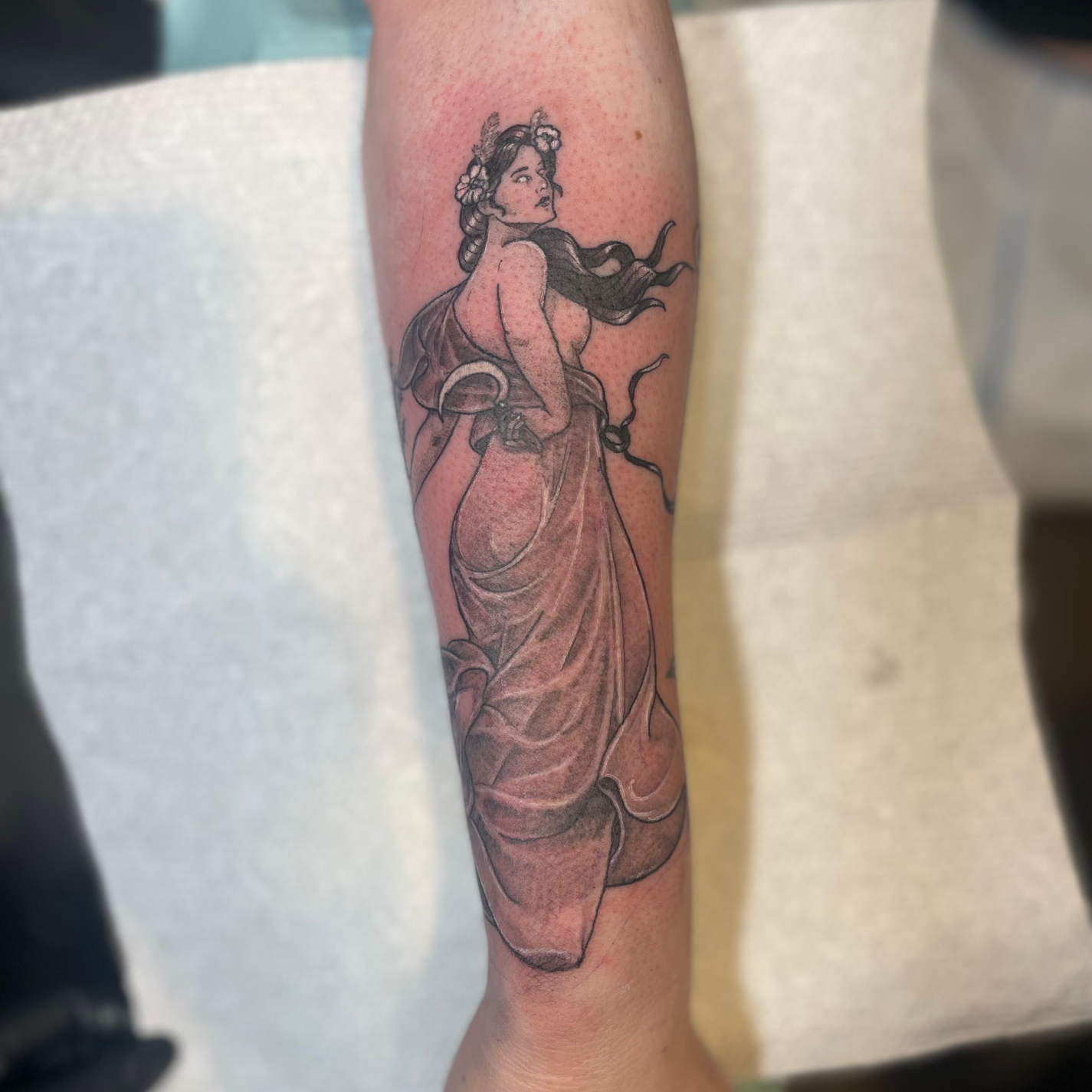 Tattoo on forearm of a woman with flowing hair adorned with flowers, draped in a long, flowing garment.