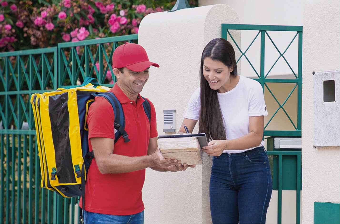 Delivery man in red uniform with large yellow backpack handing a package to a smiling woman signing on a clipboard outside a house.