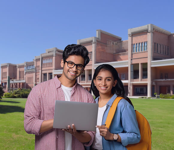 Smiling male and female students holding a laptop on a college campus lawn with academic buildings in the background.
