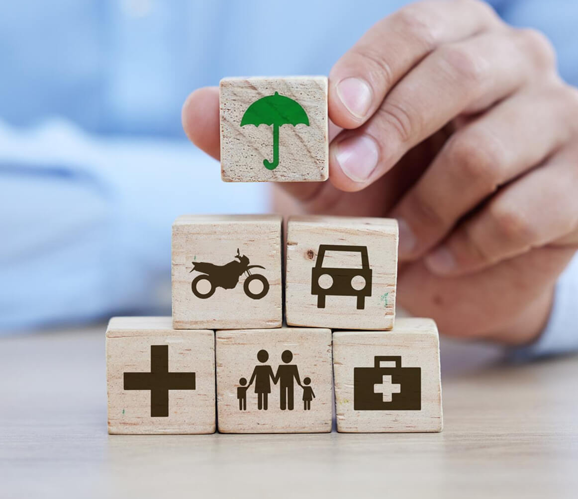 Wooden blocks with icons of motorcycle, car, family, medical cross, medical kit, topped by a block with a green umbrella symbol.