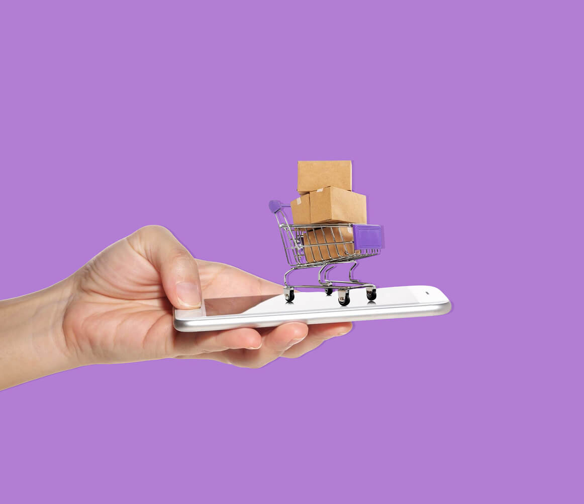 Hand holding a smartphone with a miniature shopping cart filled with cardboard boxes on the screen, symbolizing online shopping.