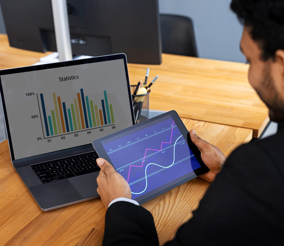 Man in business attire holding a tablet showing line graphs, with a laptop displaying colorful bar charts on a wooden desk.