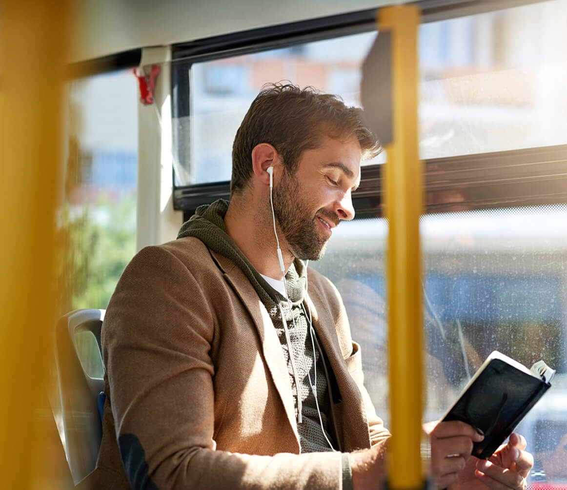 Smiling man with earphones reading a book while sitting on a bus.