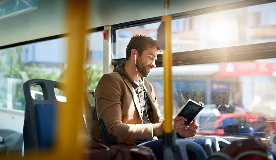 Smiling man with earphones reading a book while sitting on a bus.