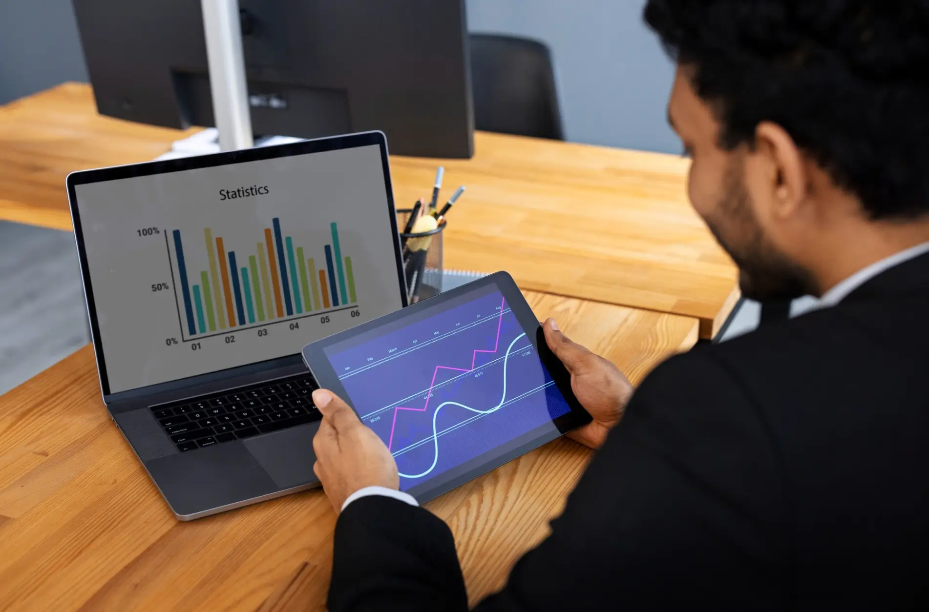 Man in business attire holding a tablet showing line graphs, with a laptop displaying colorful bar charts on a wooden desk.