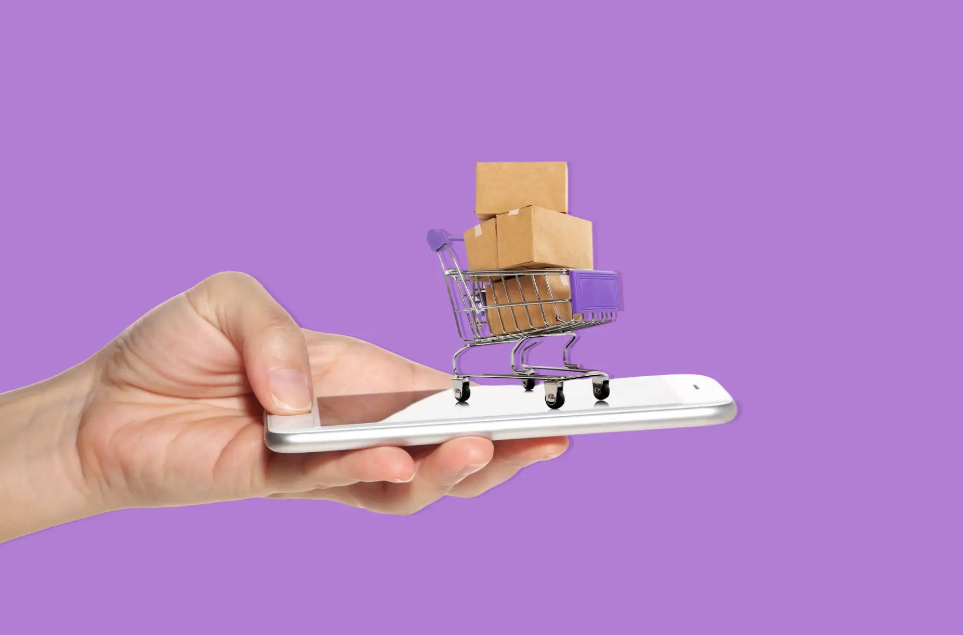 Hand holding a smartphone with a miniature shopping cart filled with cardboard boxes on the screen, symbolizing online shopping.