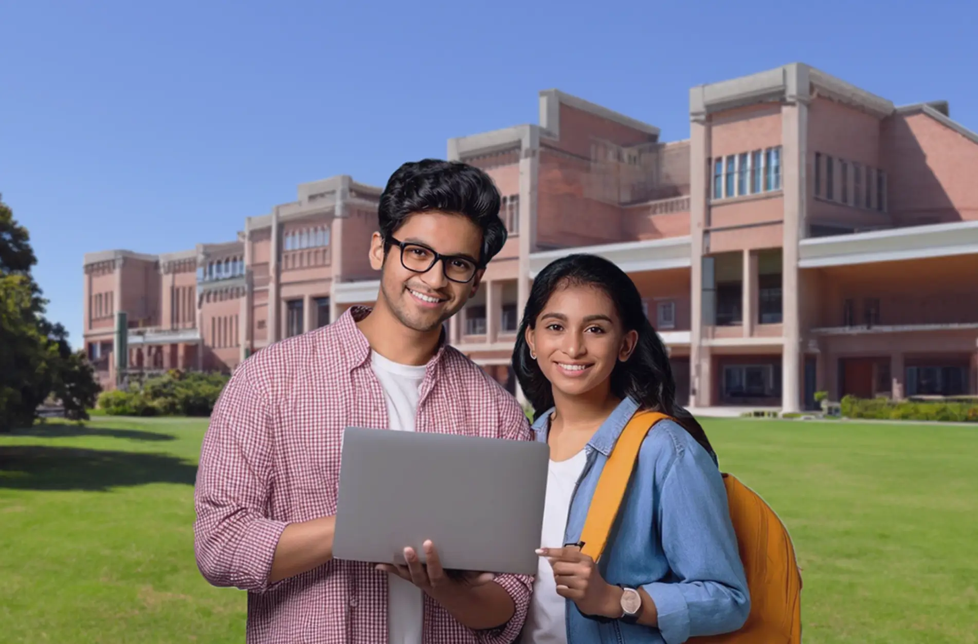 Smiling male and female students holding a laptop on a college campus lawn with academic buildings in the background.