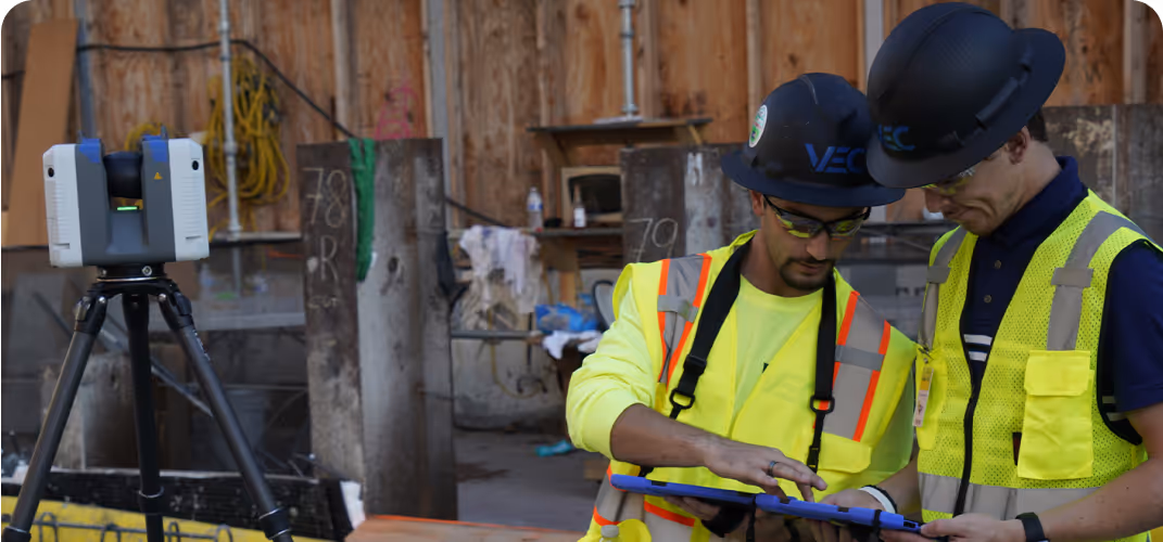 Two construction workers in safety vests and helmets review BIM data on a tablet onsite, with a tripod-mounted scanner nearby and construction materials in the background to support quality control