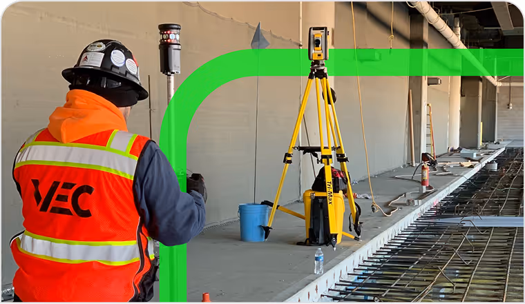A construction worker in a VEC safety vest uses surveying equipment, including a tripod and 3D laser tool, at an indoor construction site, ensuring predictable project execution.