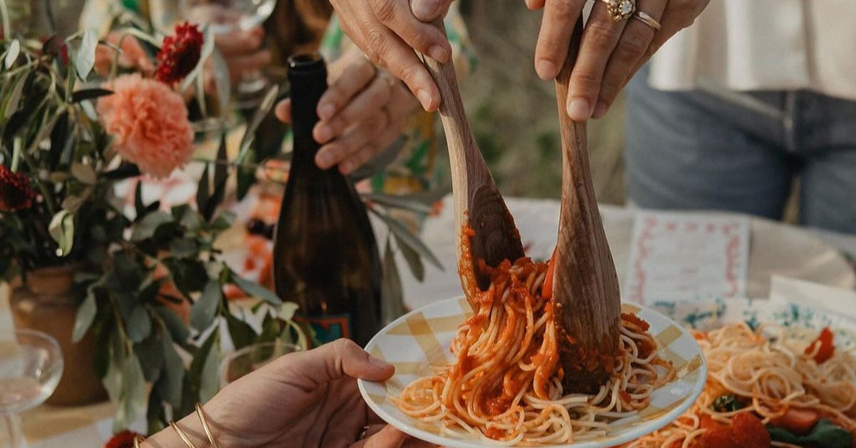 Son las manos de una mujer sirviendo spaguetti a la bolognesa con unas cucharas de madera a sus invitados en una tarde cálida