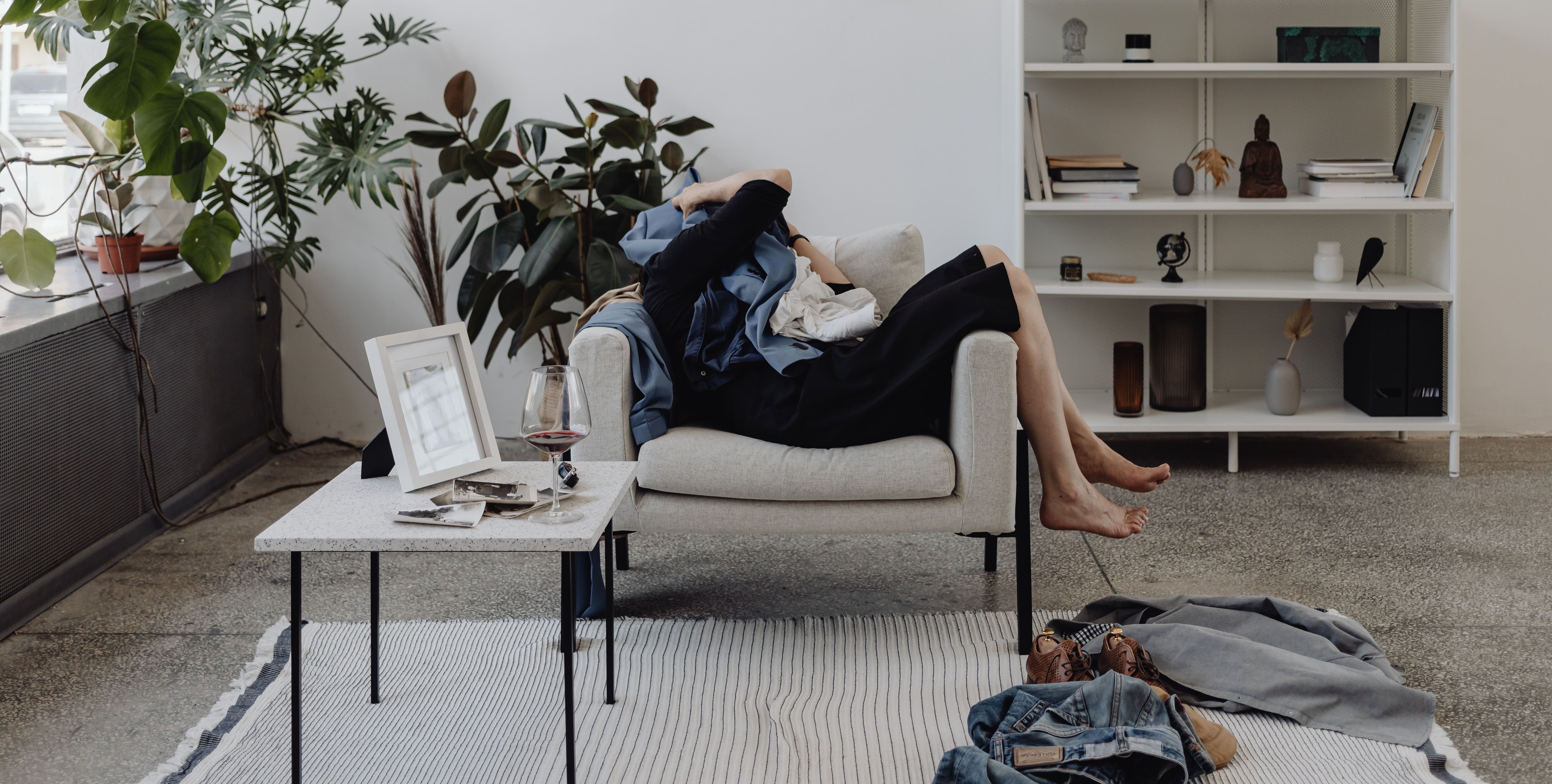 Chica sentada en su sala contemporánea monocromática con plantas de fondo, un mueble y ropa sucia en el suelo